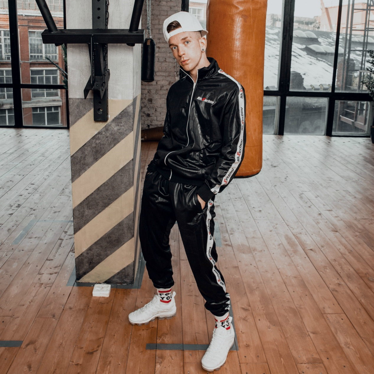 Junger Typ in einem Loft trägt einen Shiny Jogginganzug von Sk8erboy mit Basecap und nike sneaker sehr lässig